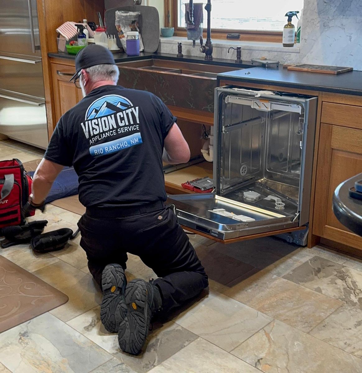 Technician kneeling on the kitchen floor installing a stainless steel dishwasher under the counter, wearing a black Vision City shirt.