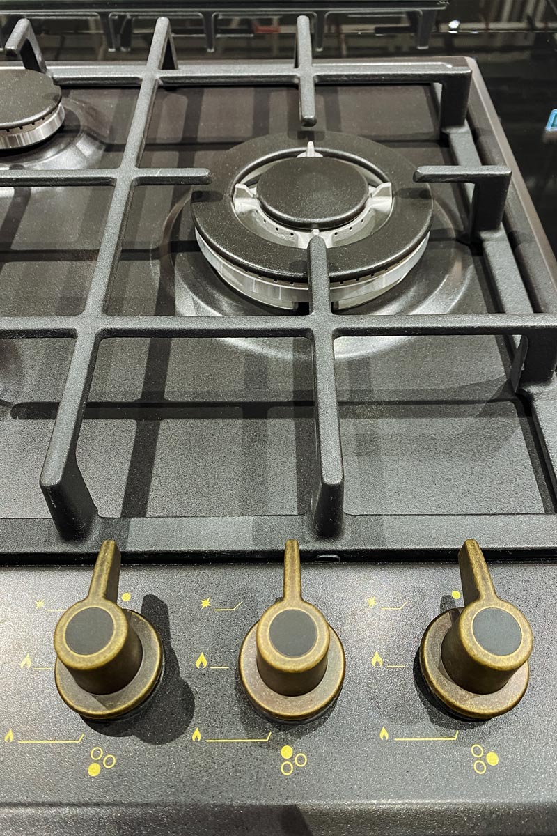 Man in a navy cap and dark T-shirt standing at a stainless steel gas stove in a kitchen, hand on the burner area.