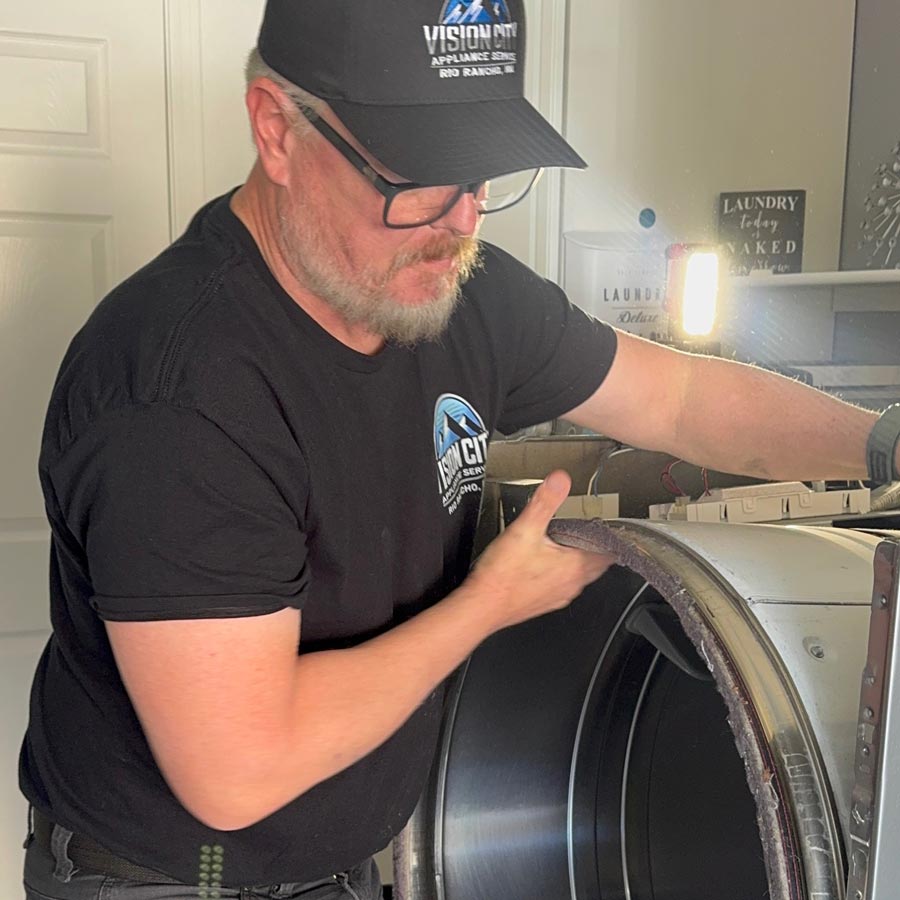 Man in a black shirt and cap using a cordless drill on top of a front-loading washer in a bright laundry room.